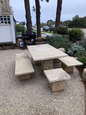 A concrete picnic table with benches in a gravel area, surrounded by trees and a building. - Apt 9: Beautiful apartment for families near a beach. (Carpinteria)