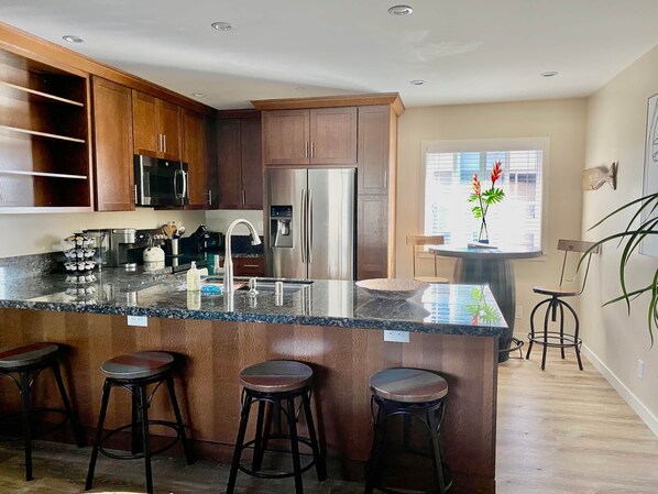 A modern kitchen with dark wood cabinets, stainless steel appliances, a granite countertop, and a central island with stools. - Apt 9: Beautiful apartment for families near a beach. (Carpinteria)