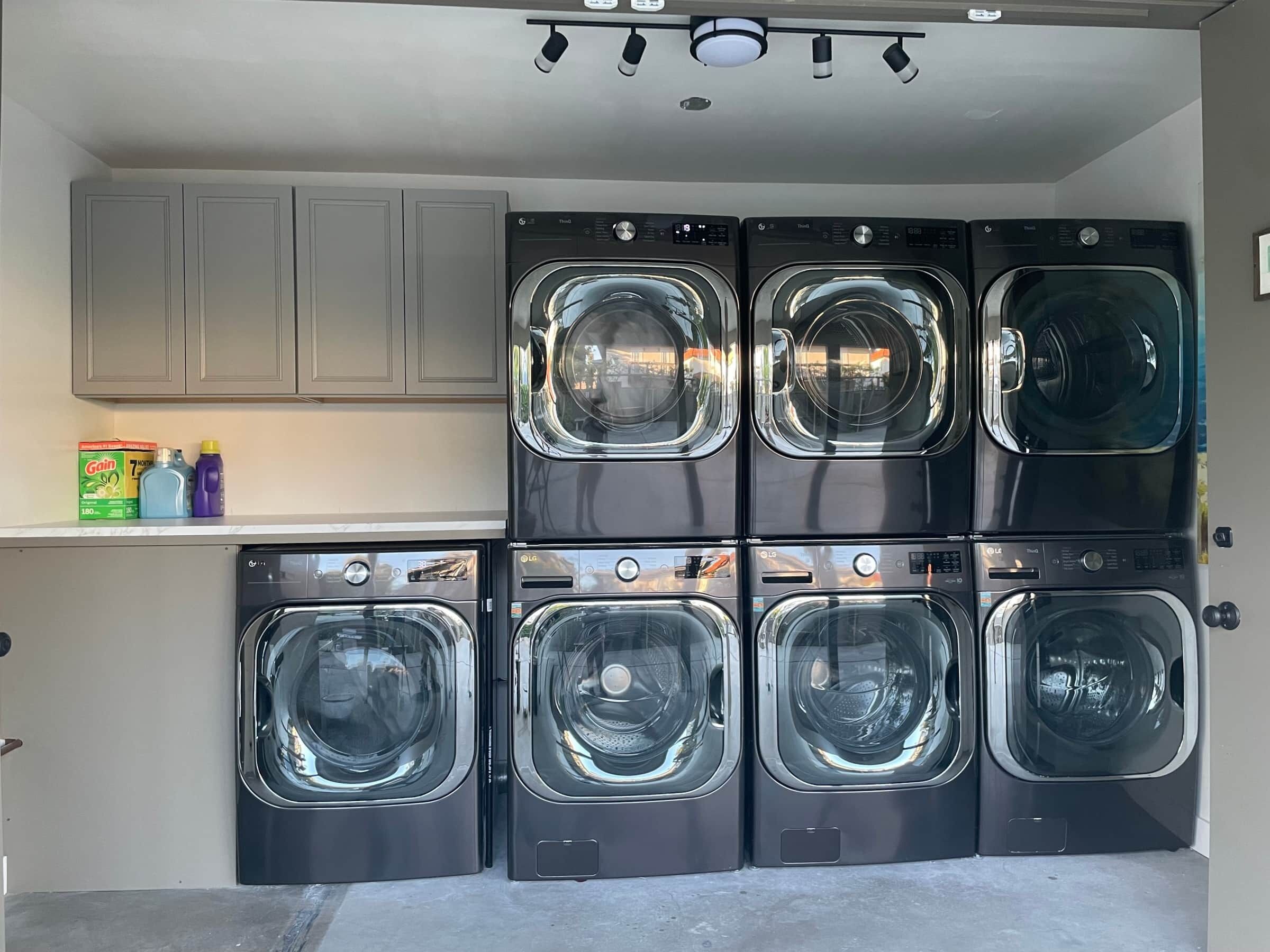 Une buanderie équipée de six machines à laver à chargement frontal superposées, d’une étagère contenant des produits de nettoyage et d’un meuble gris placé au-dessus.