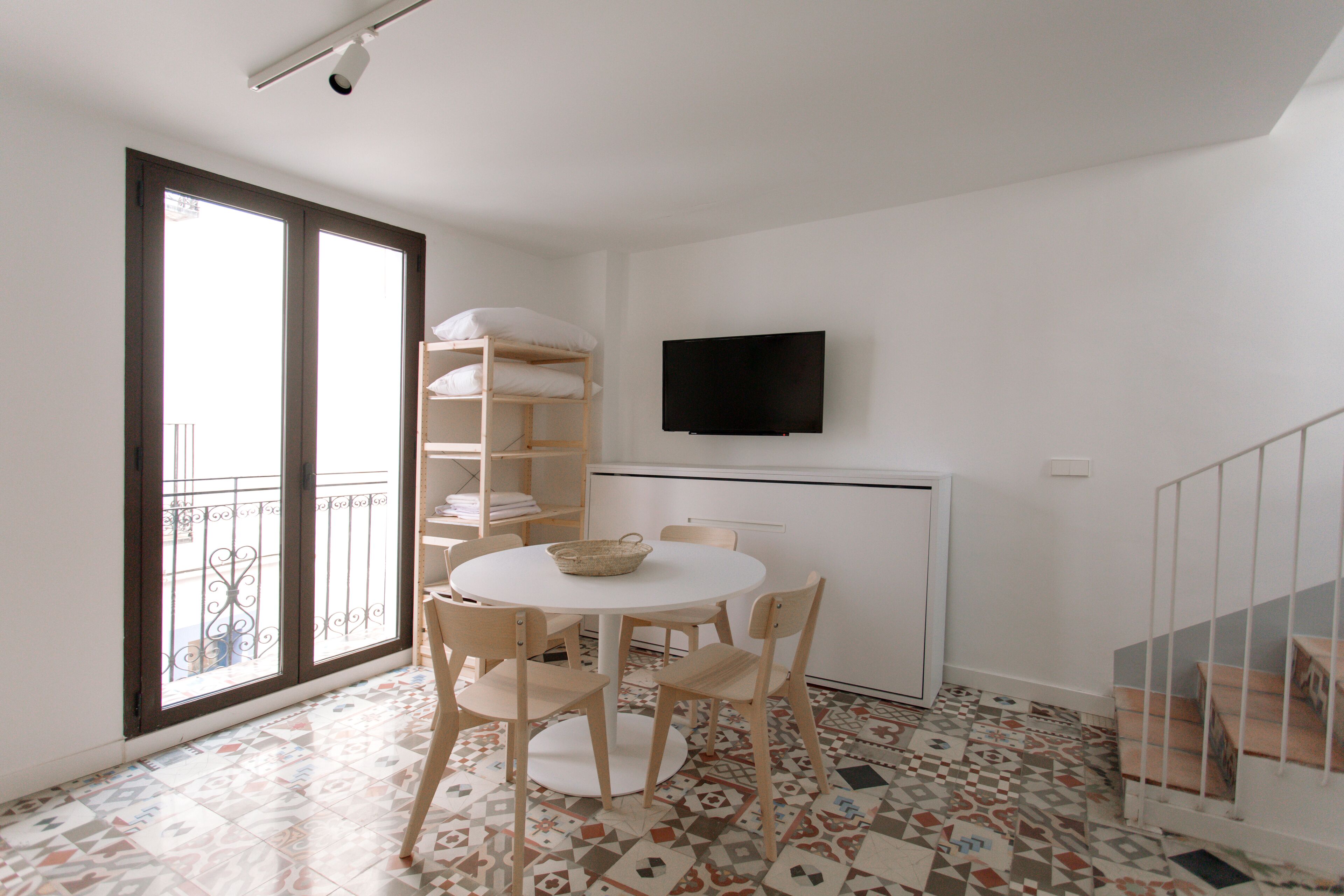 A small, bright room with a round table, chairs, a TV, a balcony with a railing, and patterned floor tiles.