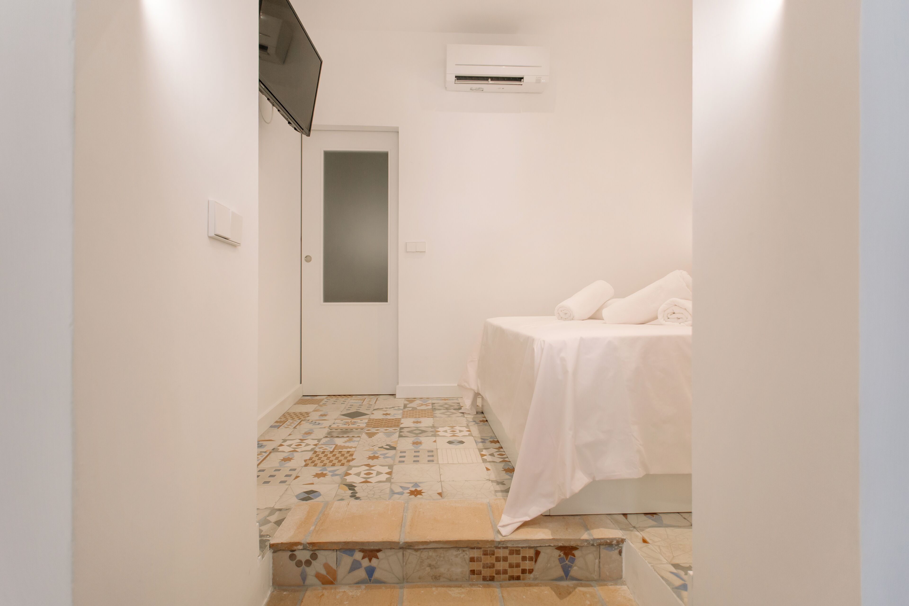 A small, clean room with a bed, a door, and a tiled floor.