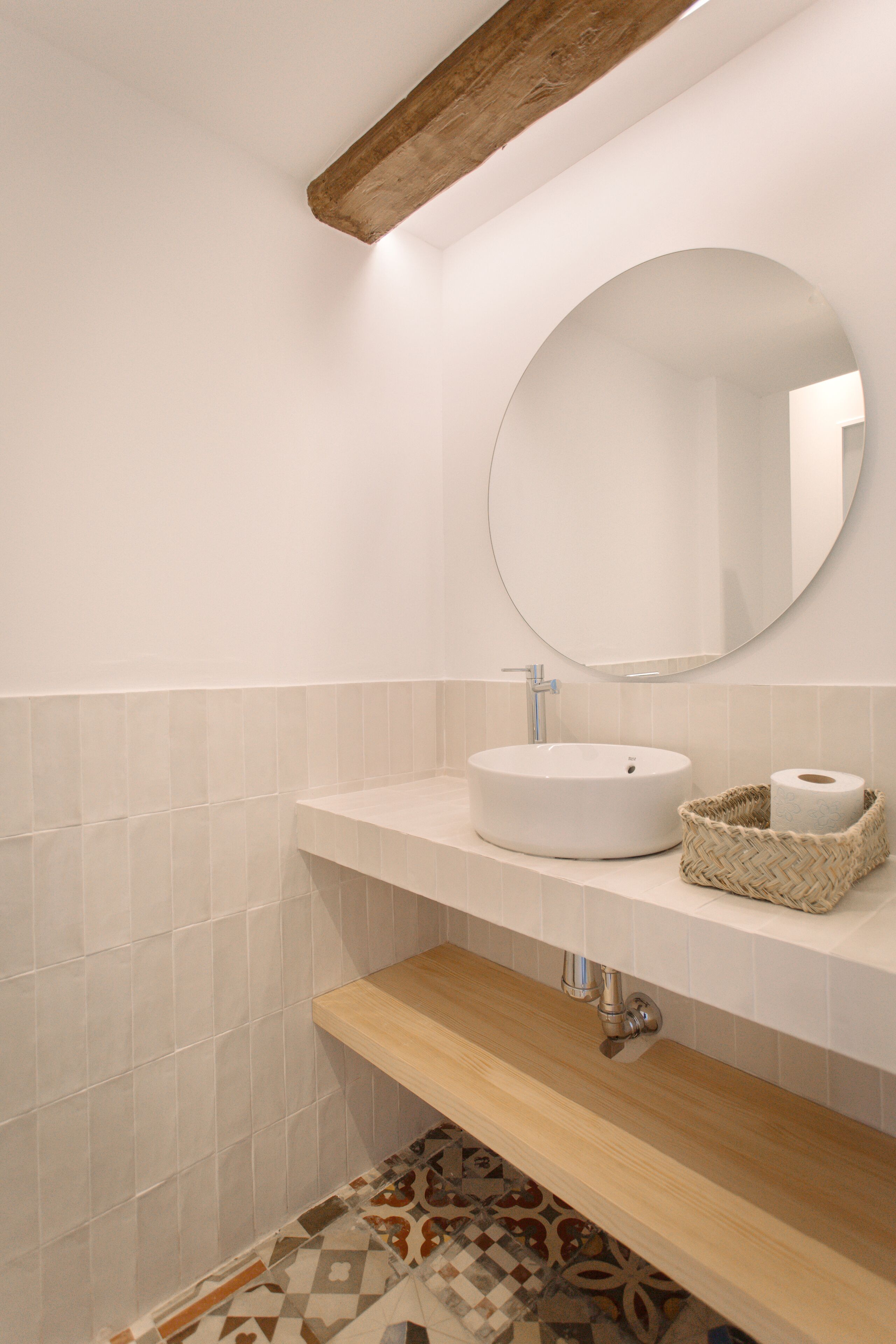 Une salle de bain avec un miroir rond, un lavabo blanc, une étagère en bois et un carrelage à motifs.