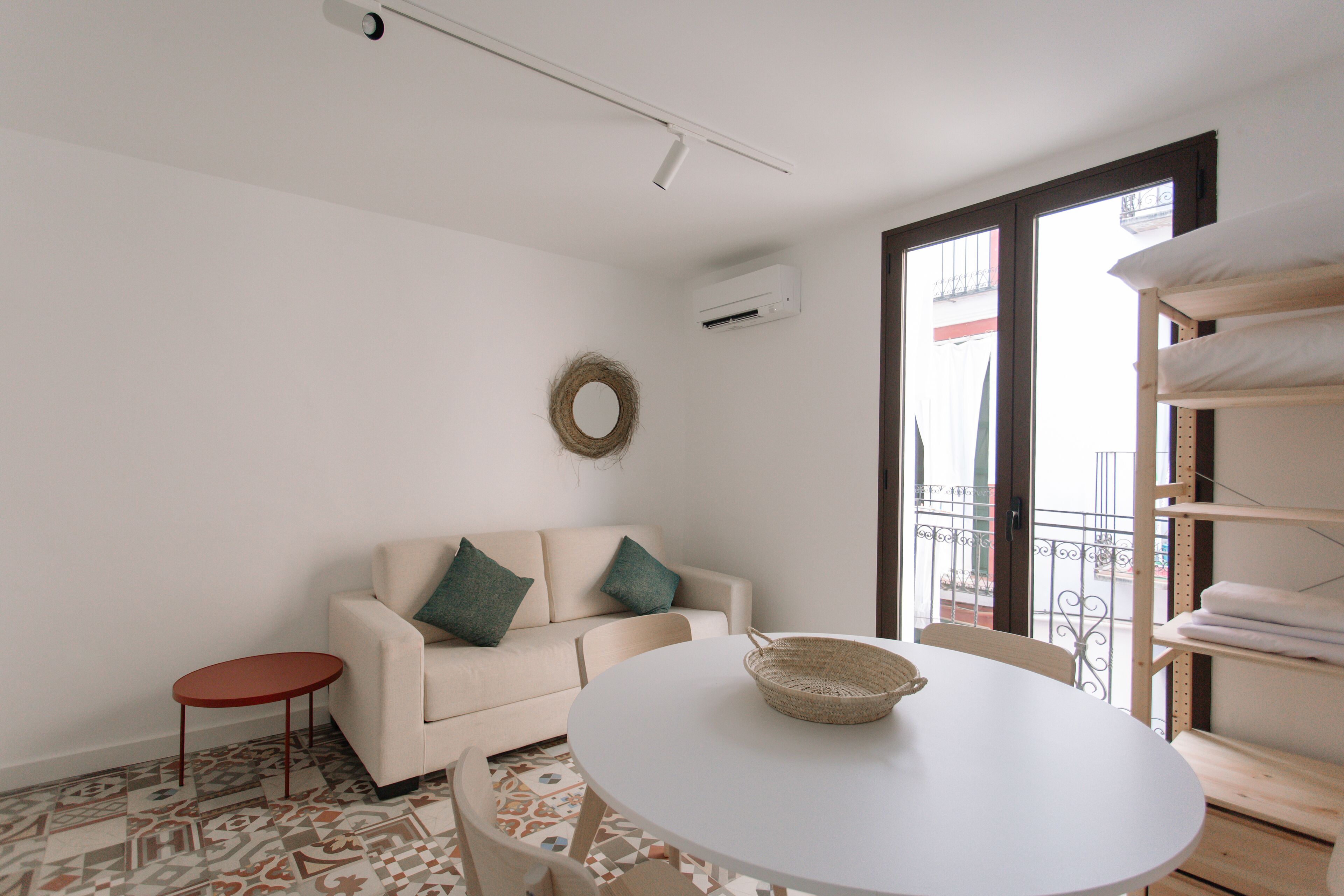 Une pièce avec une table ronde, un canapé avec des coussins, une petite table rouge, un panier en osier et une fenêtre donnant sur un balcon.