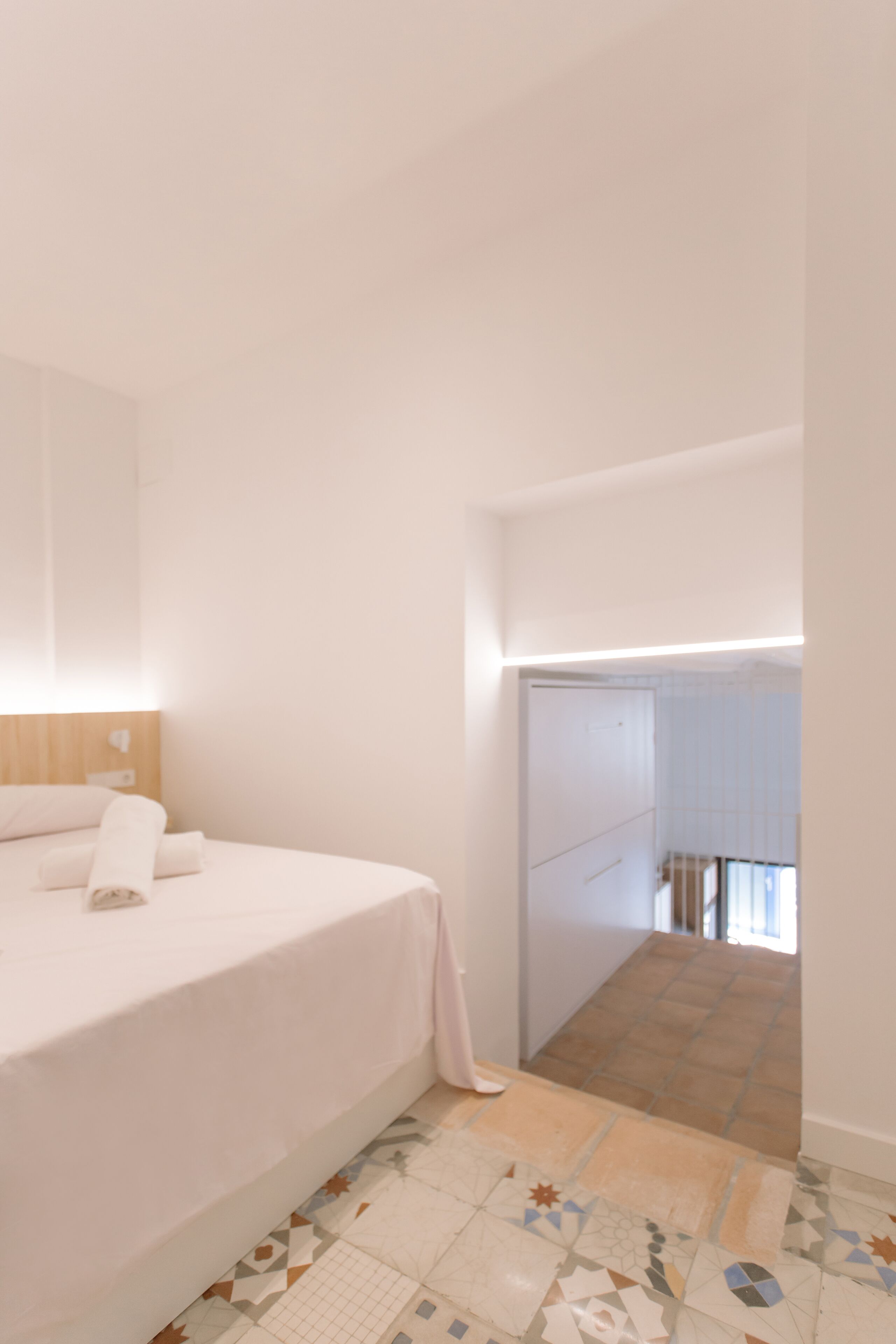 Un dormitorio con cama, mesita de noche y un espejo grande. Se ve una puerta que da a un baño y un pasillo.