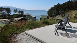 A concrete patio with a single black chair and a table, overlooking a body of water with buildings and trees in the background.