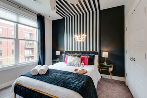 A modern hotel room with a large bed, a bedside table with a lamp, a neon sign on the wall, and a window with curtains.
