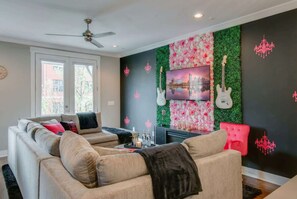 Una sala de estar moderna con un sofá seccional, un televisor de pantalla plana, guitarras decorativas y una pared verde con diseños florales.