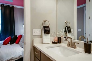 A hotel bathroom with a white sink, a modern faucet, and a mirror. There are two towels hanging on towel rings, one labeled "MAKEUP" and the other "NIGHTLIFE".
