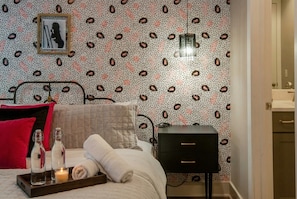 A neatly made bed with a red pillow, a bedside table with a lamp, and a tray with a candle and two bottles.
