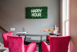 Un área de comedor con una mesa redonda, cuatro sillas rosas, un letrero de neón que dice 'HAPPY HOUR' y un letrero que dice 'COFFEE TIME'.