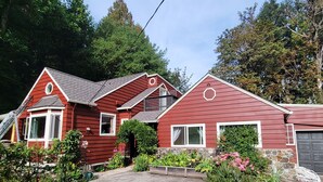 A red two-story house with a stone foundation, a garage, and a front yard with various plants and flowers. - All the amenities of home with no cleaning fees and first breakfast included!  (West Linn)