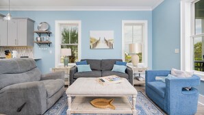 Una sala de estar con un sofá gris, un sillón azul y un reposapiés blanco. Hay un cuadro en la pared y una ventana con vista a follaje.