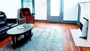 Ein Wohnzimmer mit einem Ledersofa, einem roten Sessel, einem Couchtisch und einem gemusterten Teppich. Im Hintergrund ist eine Tür und ein weißer Heizkörper zu sehen.