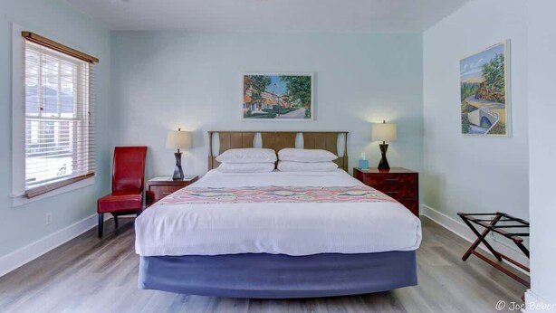 Une chambre à coucher avec un lit, deux tables de chevet, une chaise rouge, un ventilateur de plafond et deux tableaux accrochés aux murs.