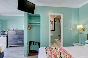 A bedroom with teal walls, a bed with a colorful blanket, a TV mounted on the wall, a dresser, and a bathroom visible through an open door. - Unit 11 Dbl Queen Room, Mini Fridge, Microwave, Wi (Hot Springs)
