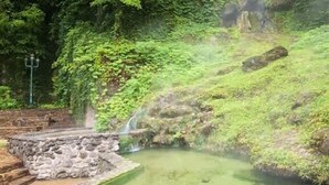 Eine natürliche Thermalquellenlandschaft mit einem steinernen Pfad, der zu einem kleinen Becken inmitten üppiger Vegetation und einem Wasserfall führt.