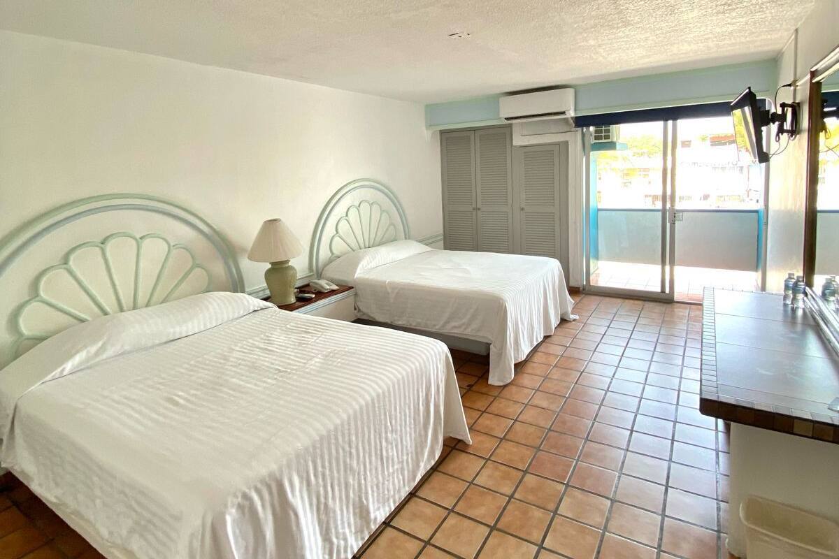 A hotel room with two beds, a small desk, a chair, a window with blinds, and a tiled floor.