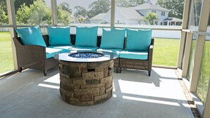 A covered patio area with wicker furniture and teal cushions, a stone fire pit, and a view of a residential neighborhood.