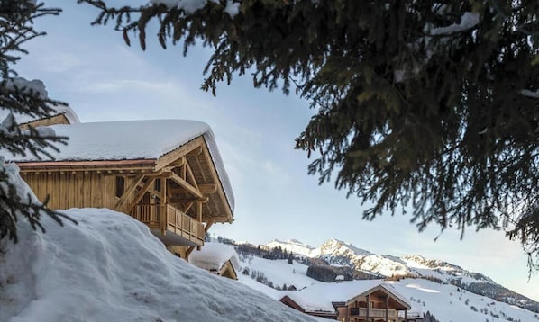 Una cabaña de madera con techo nevado, rodeada de montañas y árboles cubiertos de nieve.