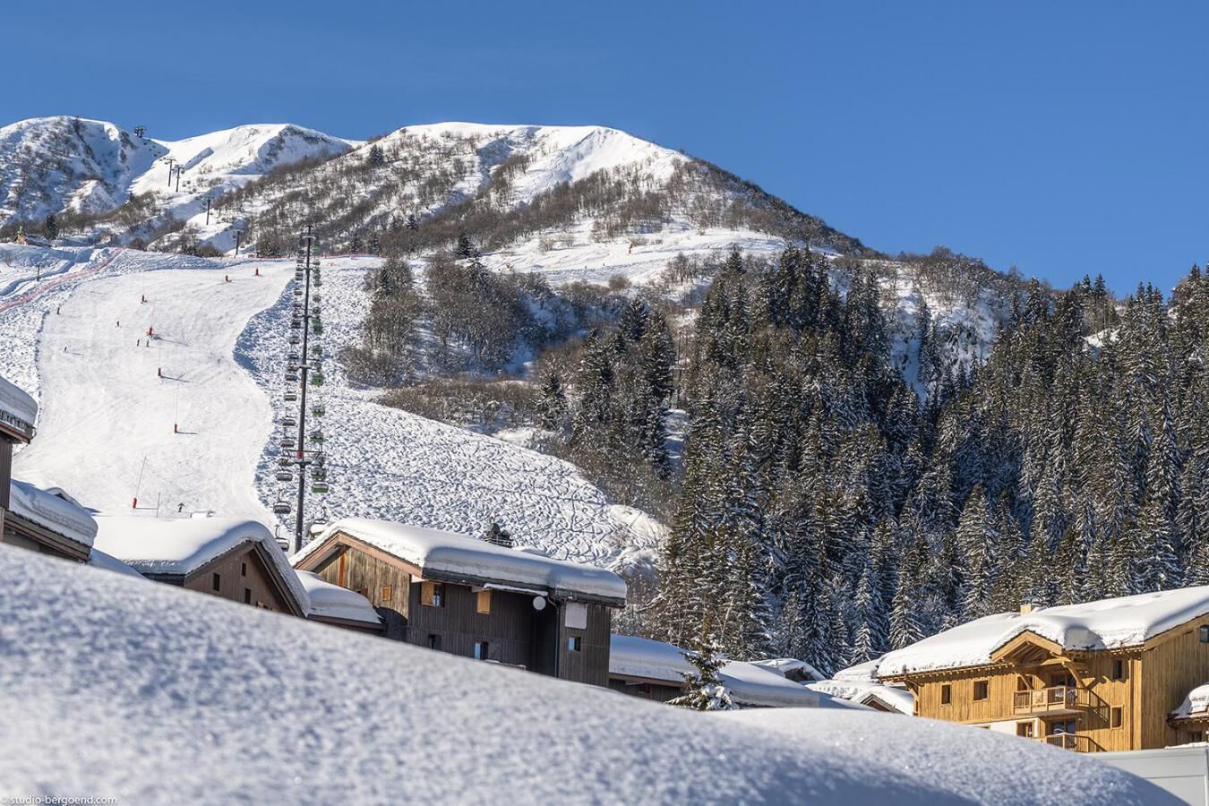 A snow-covered mountain with a ski slope, wooden buildings, and a clear blue sky.