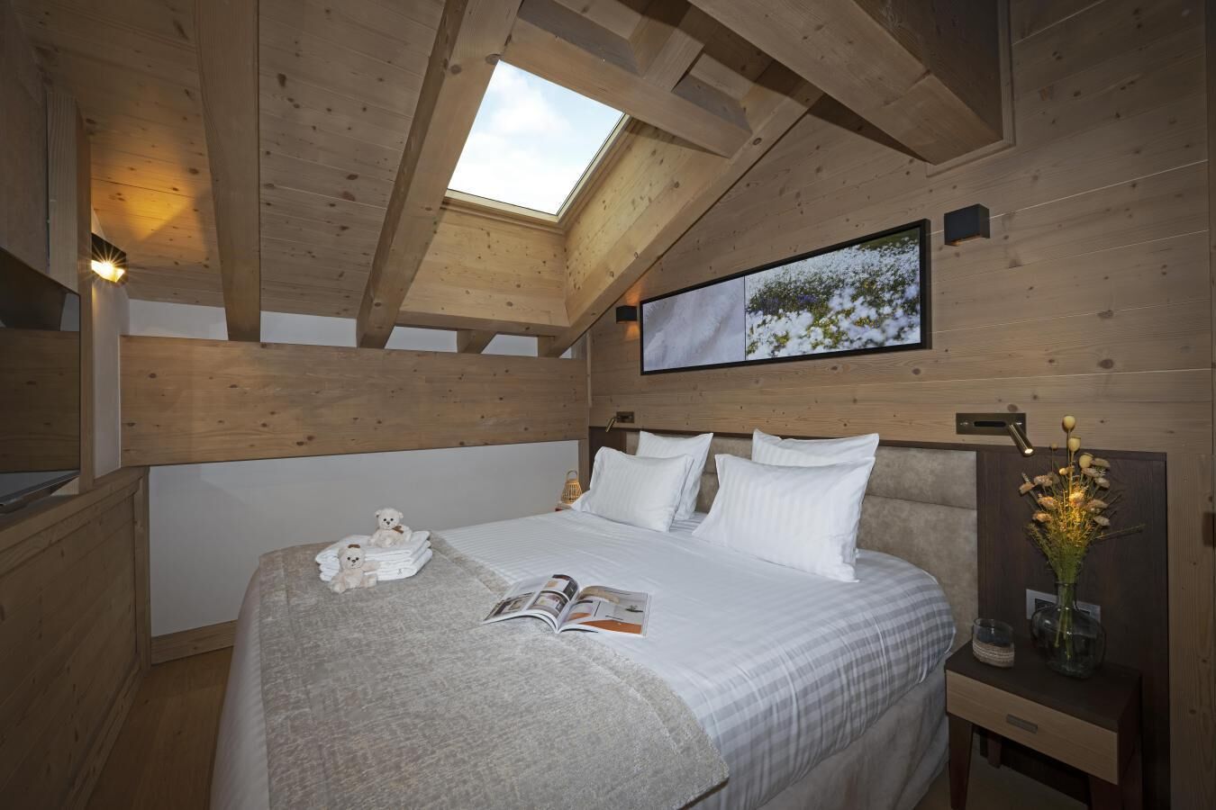 A cozy bedroom with a wooden ceiling, a skylight, a bed with white linens, a bedside table with a lamp, and a framed picture on the wall.