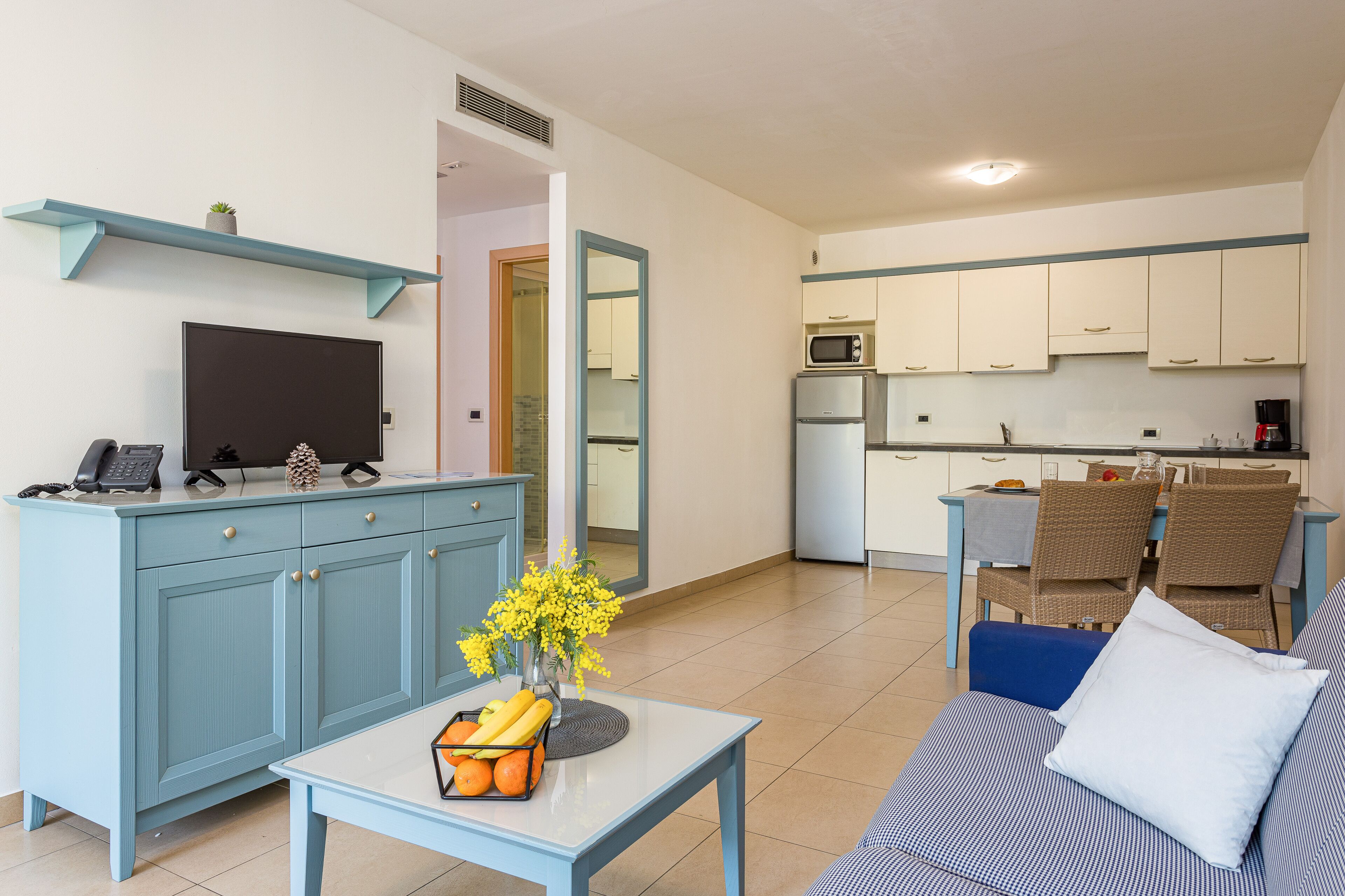 Una sala de estar moderna con un sofá, una mesa de centro y un televisor sobre un mueble bajo. Se ve una cocina al fondo, con gabinetes blancos y una zona de comedor con sillas de mimbre.