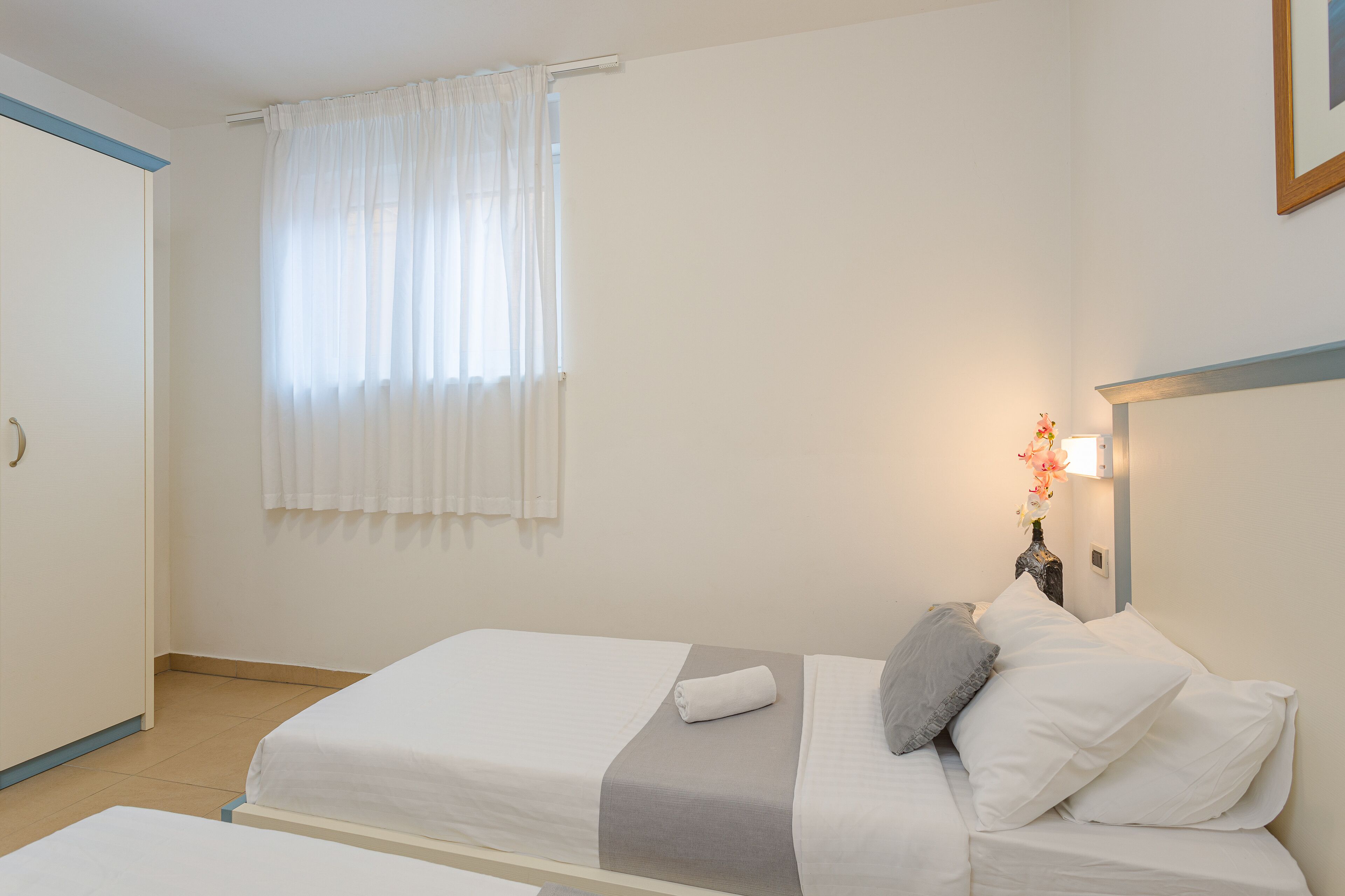 Una camera d'albergo con due letti, un comodino con un vaso di fiori e una finestra con tende leggere.
