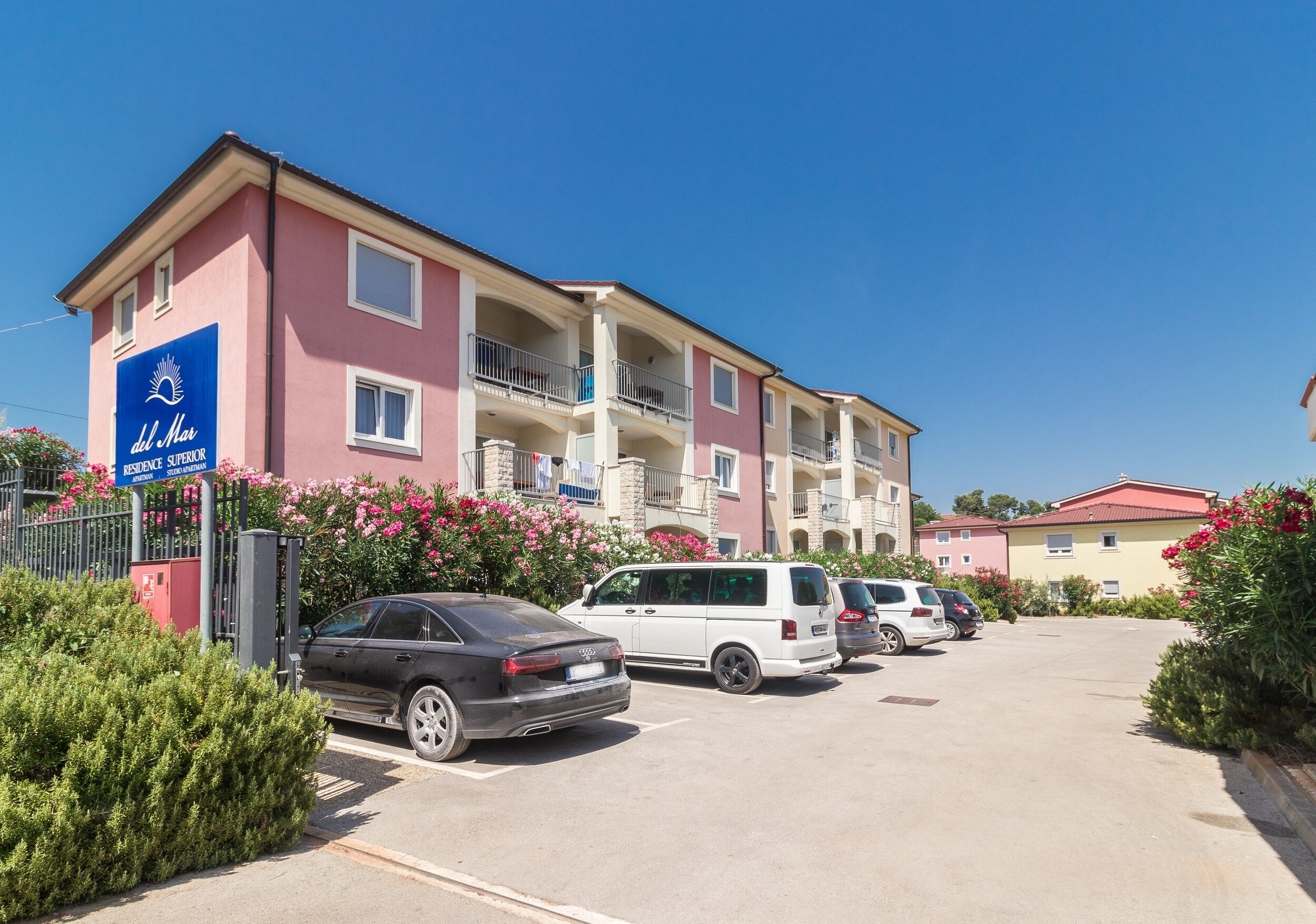 A multi-story hotel with a pink facade, balconies, and a parking area with several cars.