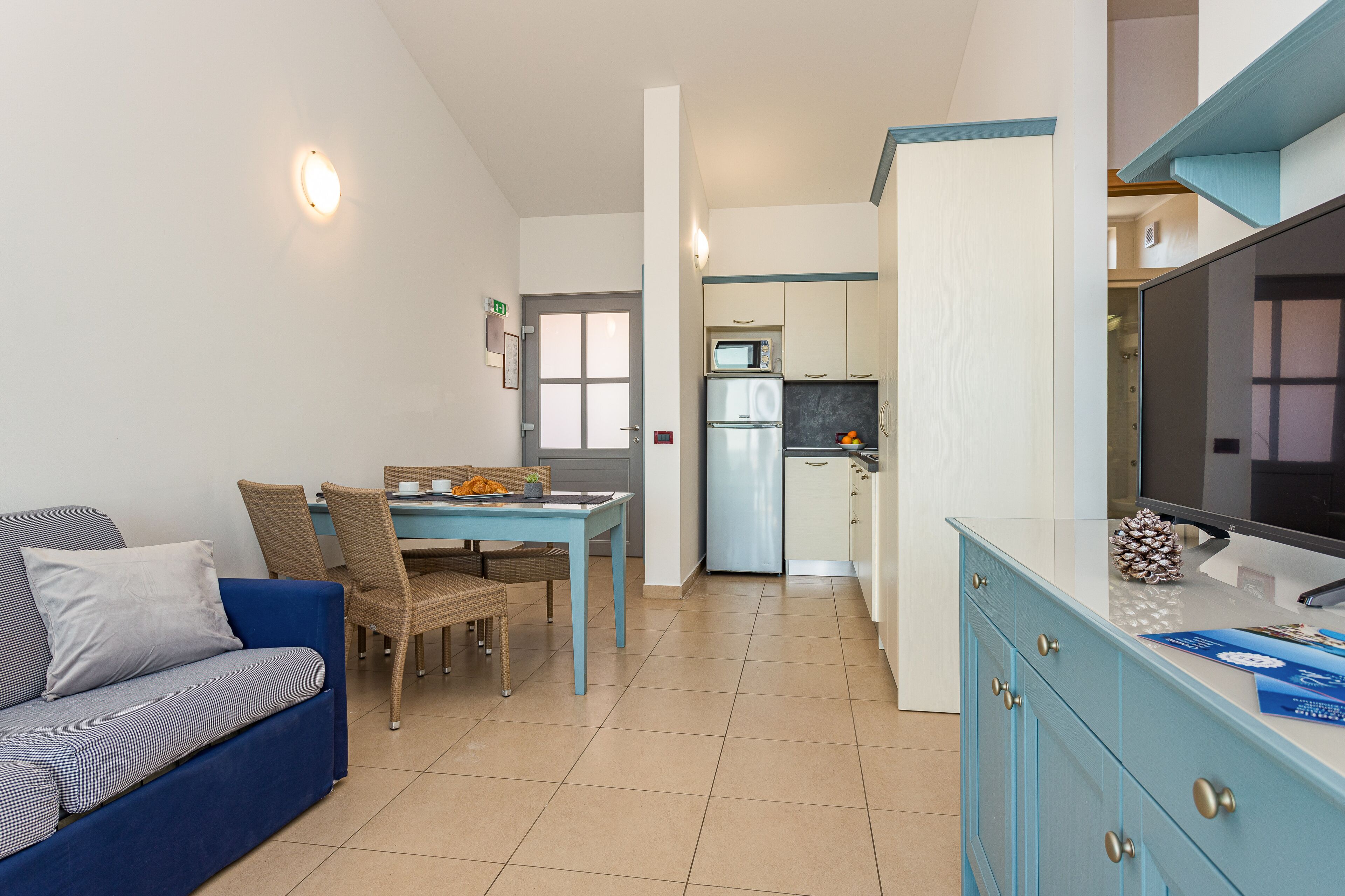A compact living space with a sofa, dining table, chairs, a kitchen area with a microwave and refrigerator, and a TV on a cabinet.