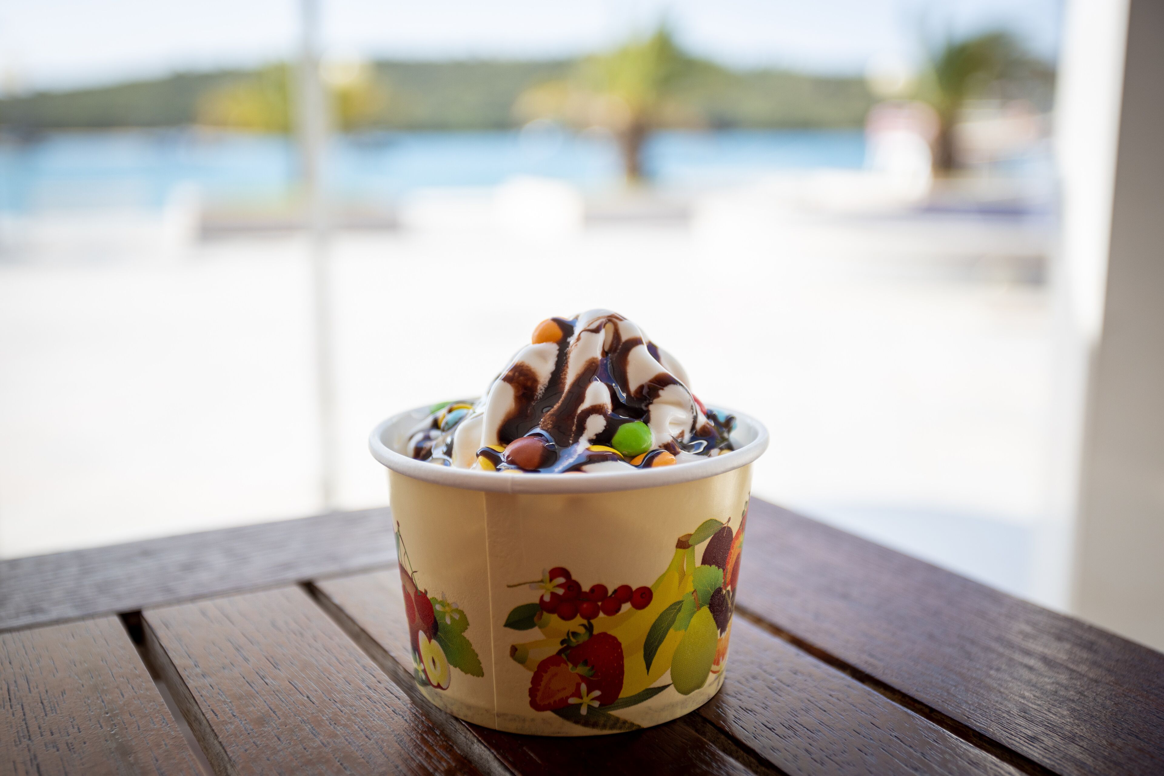 A bowl of dessert with fruit illustrations on the side, placed on a wooden table with a view of a body of water in the background.