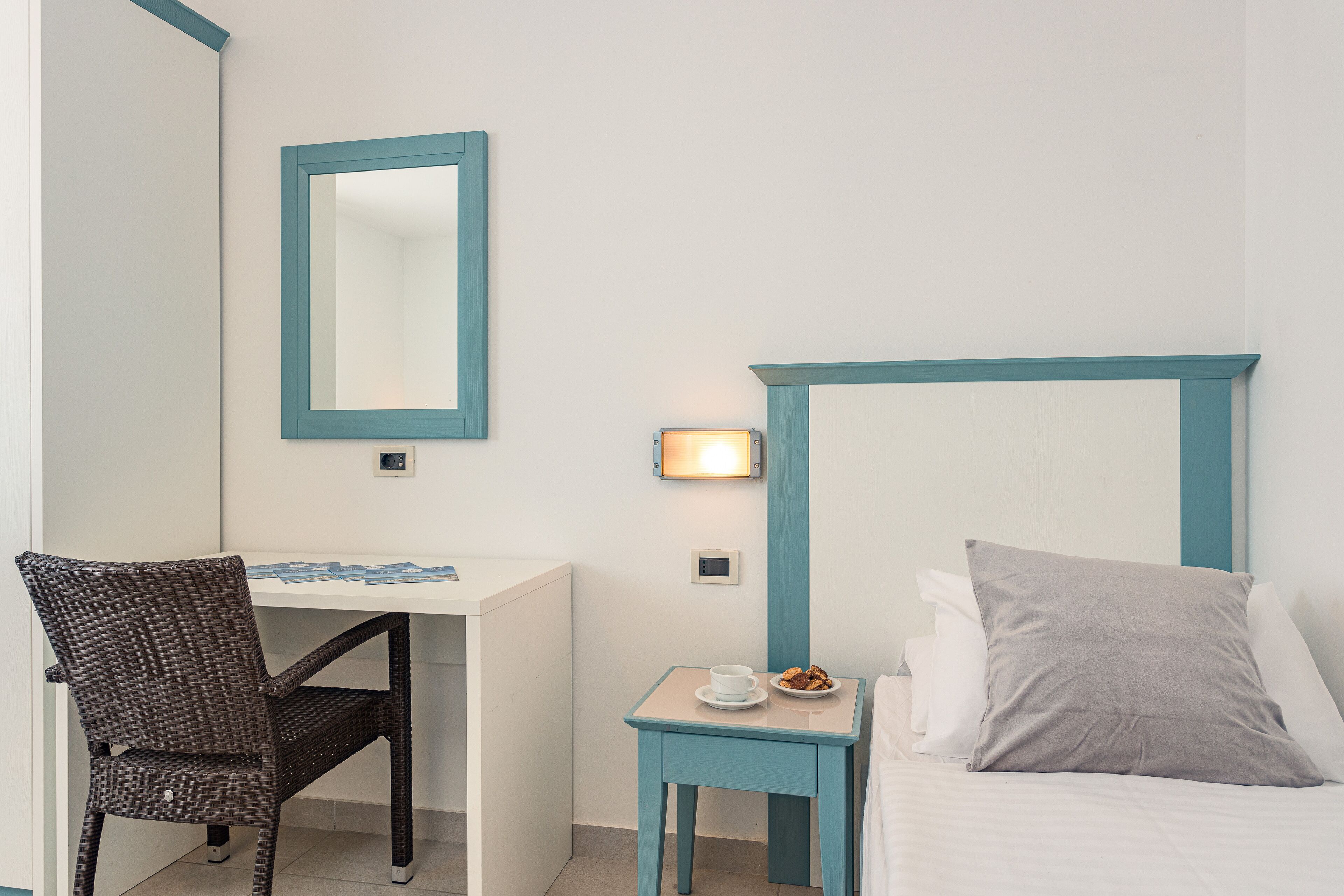 A hotel room with a bed, a desk, a chair, a small table with a cup and a plate of food, and a wall-mounted light fixture.