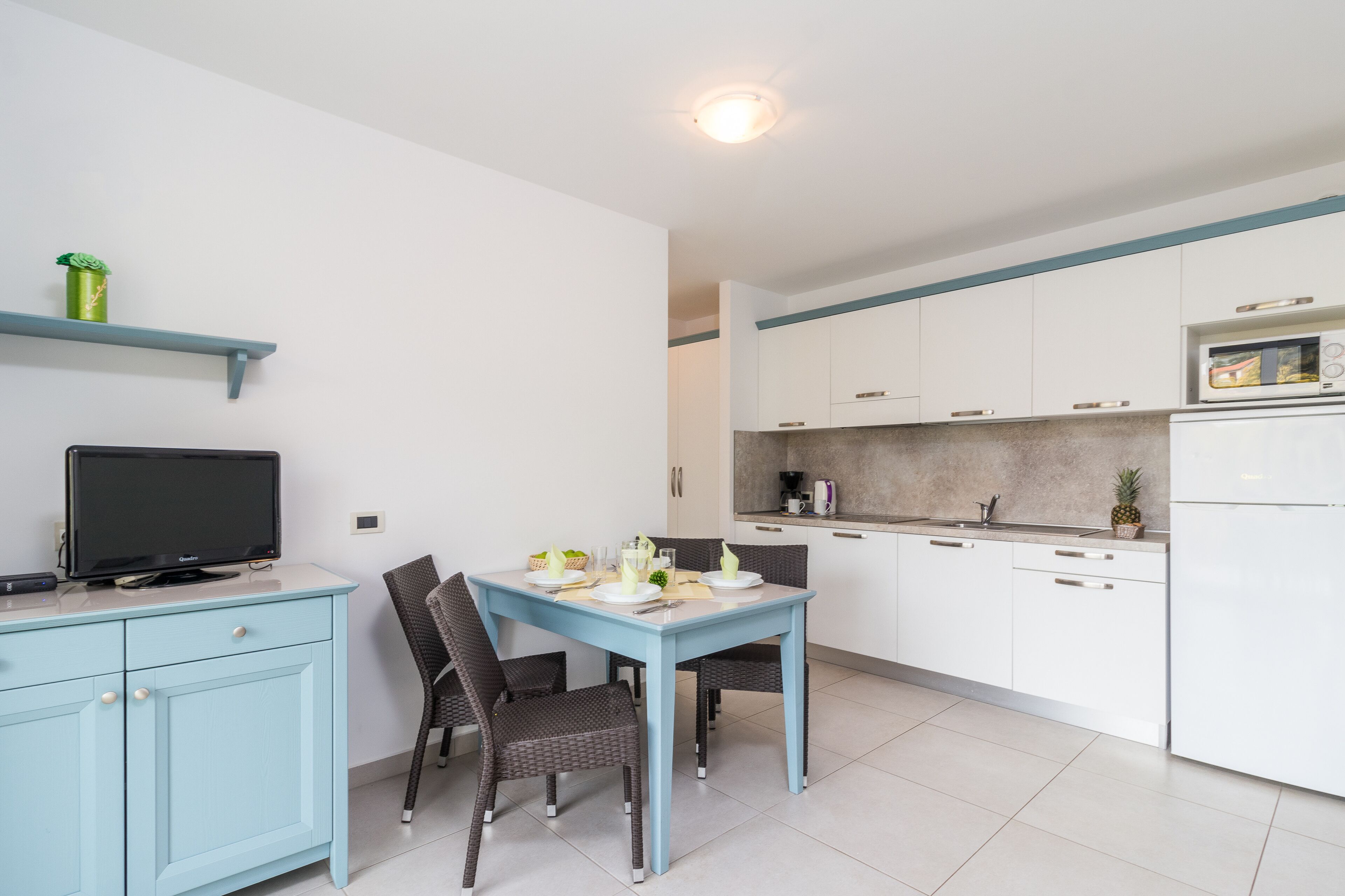 Een moderne keuken met witte kastjes, een blauwe eettafel en een flatscreen televisie op een lichtblauwe kast.