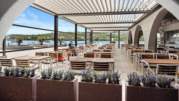An outdoor dining area with tables and chairs, large windows, and a view of the water.