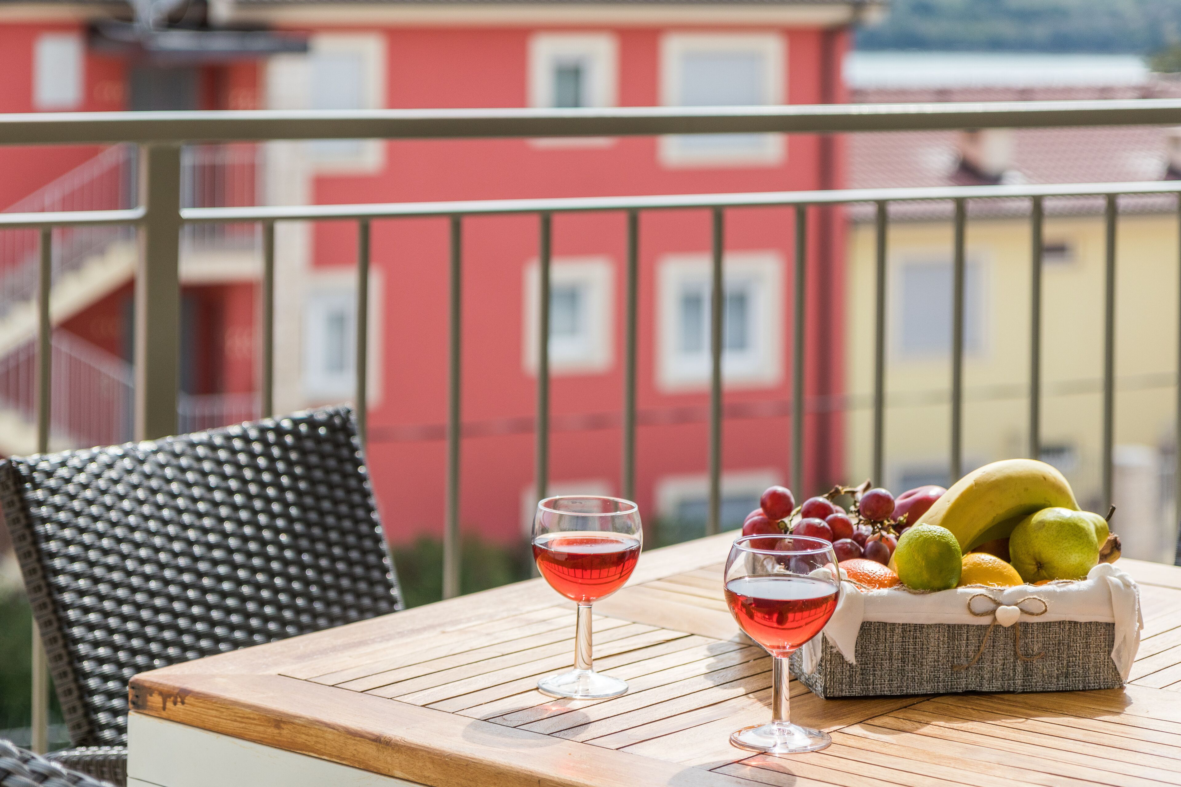 Una mesa con dos copas de vino tinto, una cesta de frutas y una silla de mimbre en un balcón con un edificio rojo al fondo.
