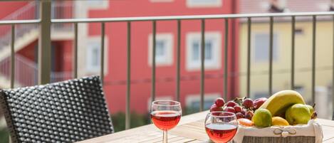 Una mesa con dos copas de vino tinto, una cesta de frutas y una silla de mimbre en un balcón con un edificio rojo al fondo.