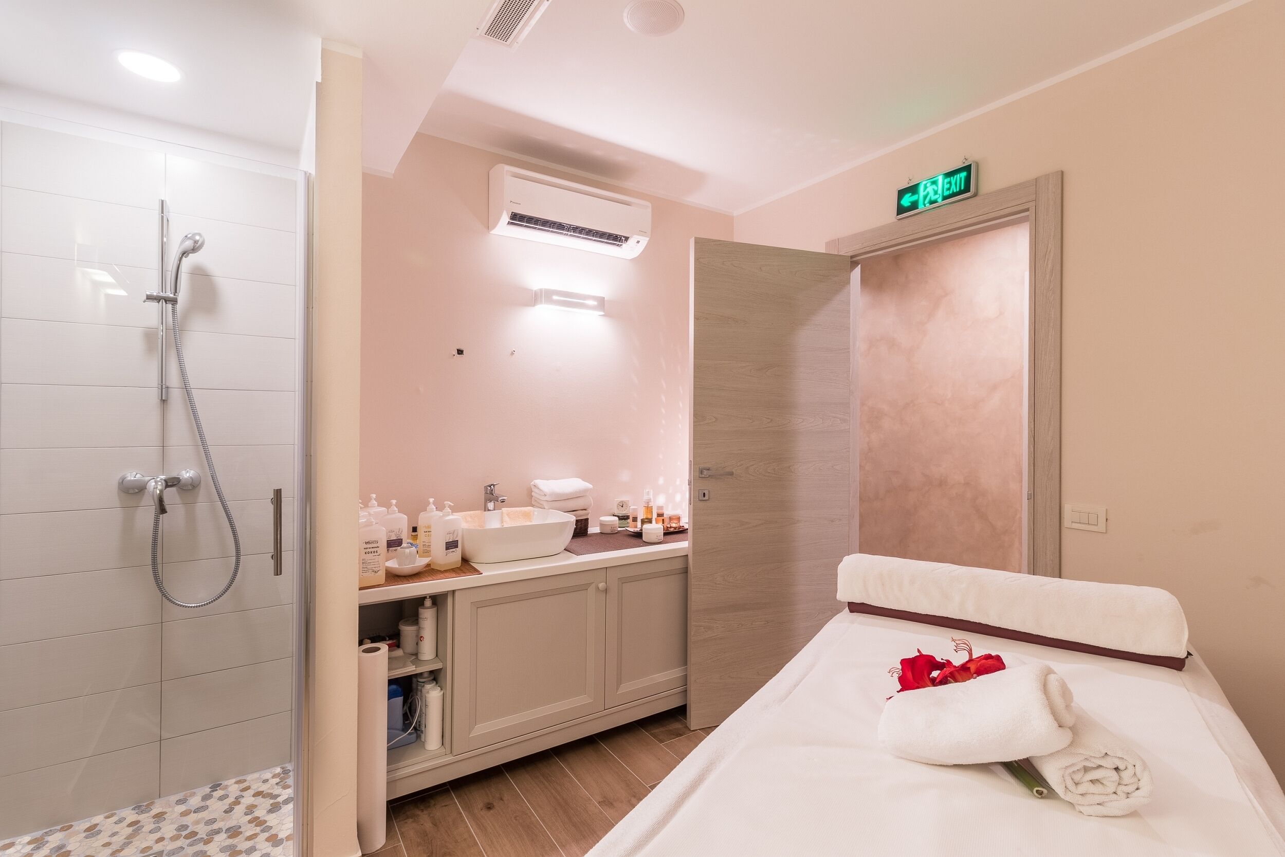 A hotel room with a shower, a sink, a bed with a towel and rose, and an exit sign.
