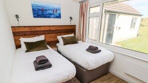 A room with two beds, a wooden headboard, a painting of sailboats, a window with curtains, and a view of a house outside. - Chalfonts Cottage (Haverfordwest)