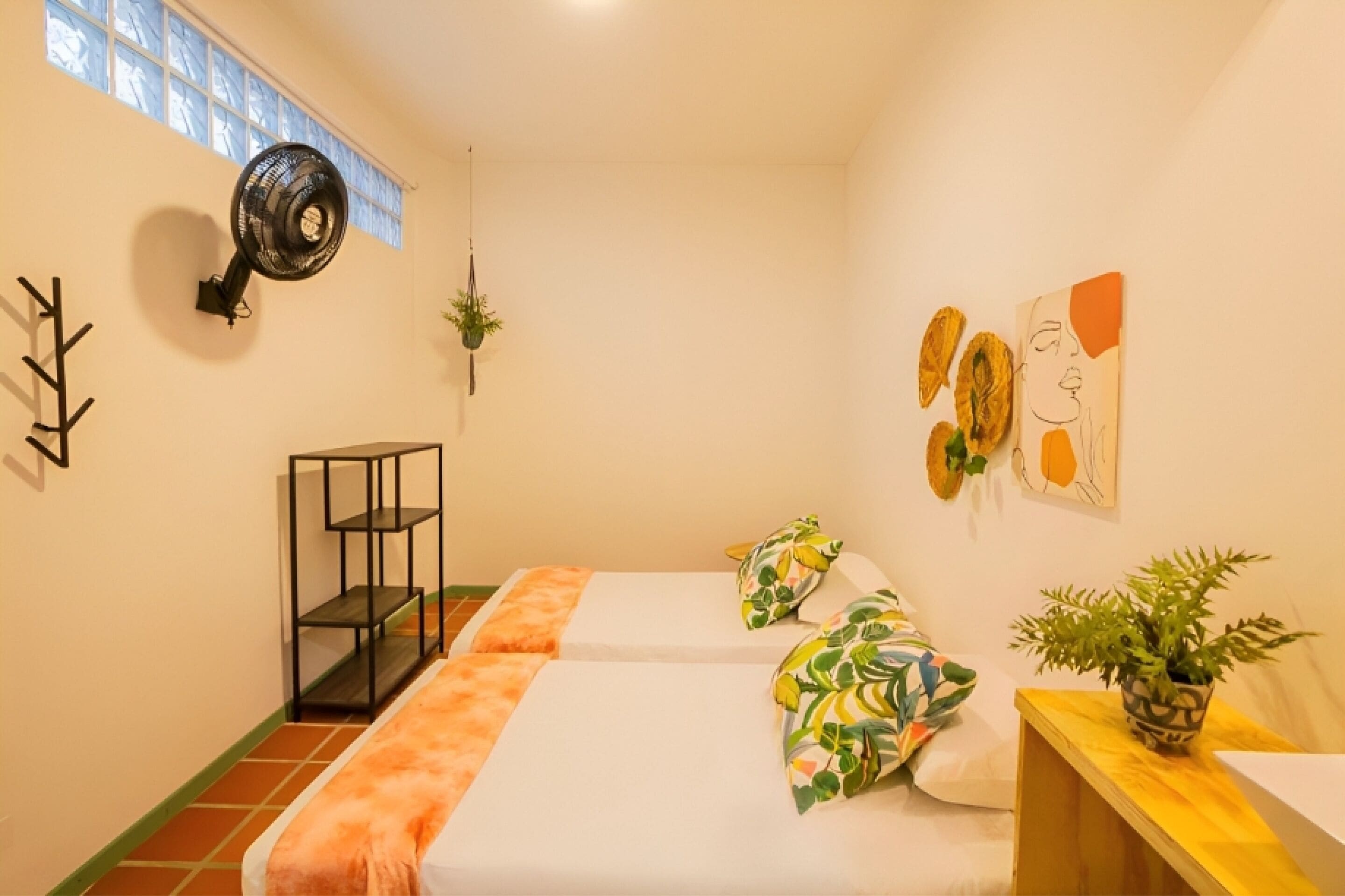 Una habitación pequeña y luminosa con dos camas, un ventilador de pie, un estante pequeño y una ventana con persianas.