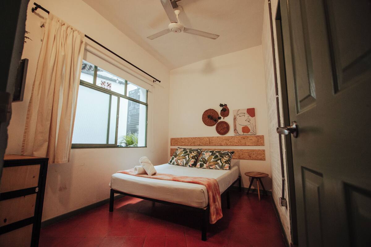 Um quarto com cama, janela com cortinas, ventilador de teto e porta.