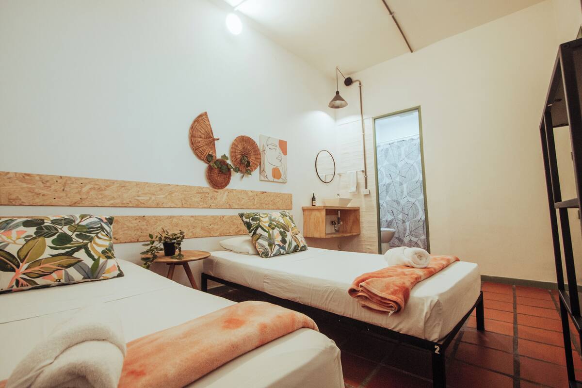 Quarto de hotel com duas camas, uma mesa pequena, uma prateleira com artigos de higiene pessoal e uma janela com vista.