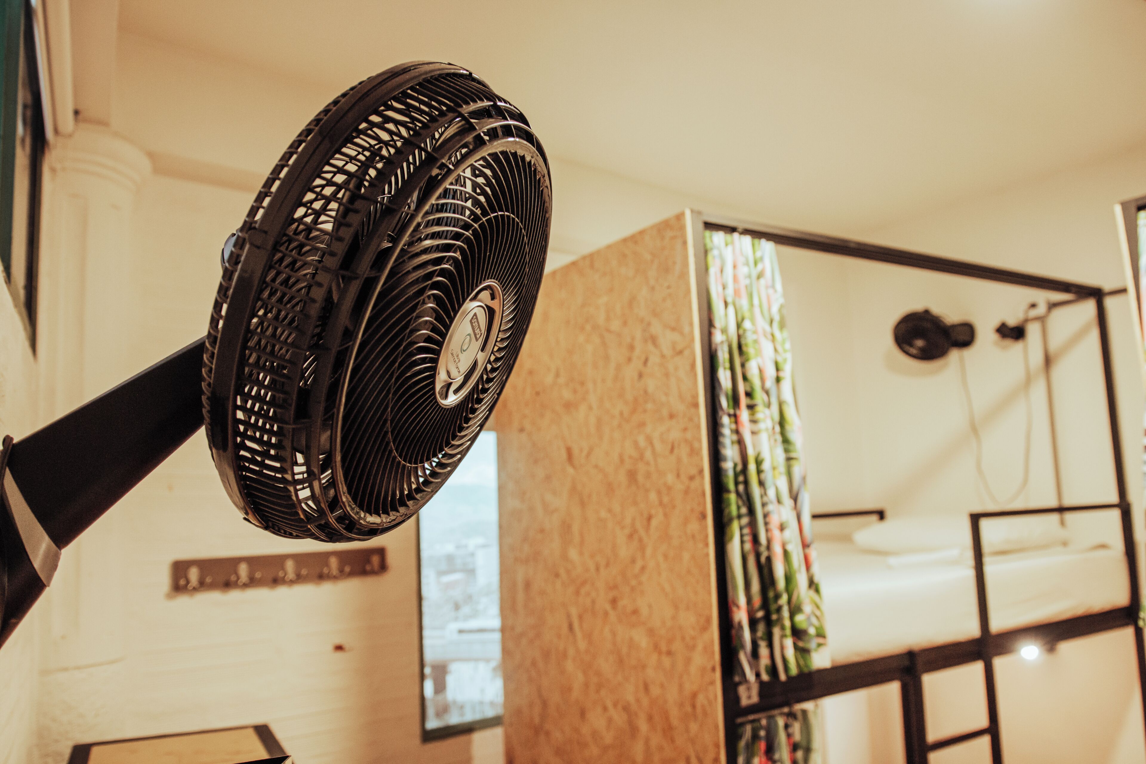 Une chambre avec un lit superposé, un ventilateur sur pied et une fenêtre avec des rideaux.
