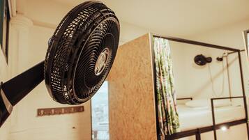 Une chambre avec un lit superposé, un ventilateur sur pied et une fenêtre avec des rideaux.