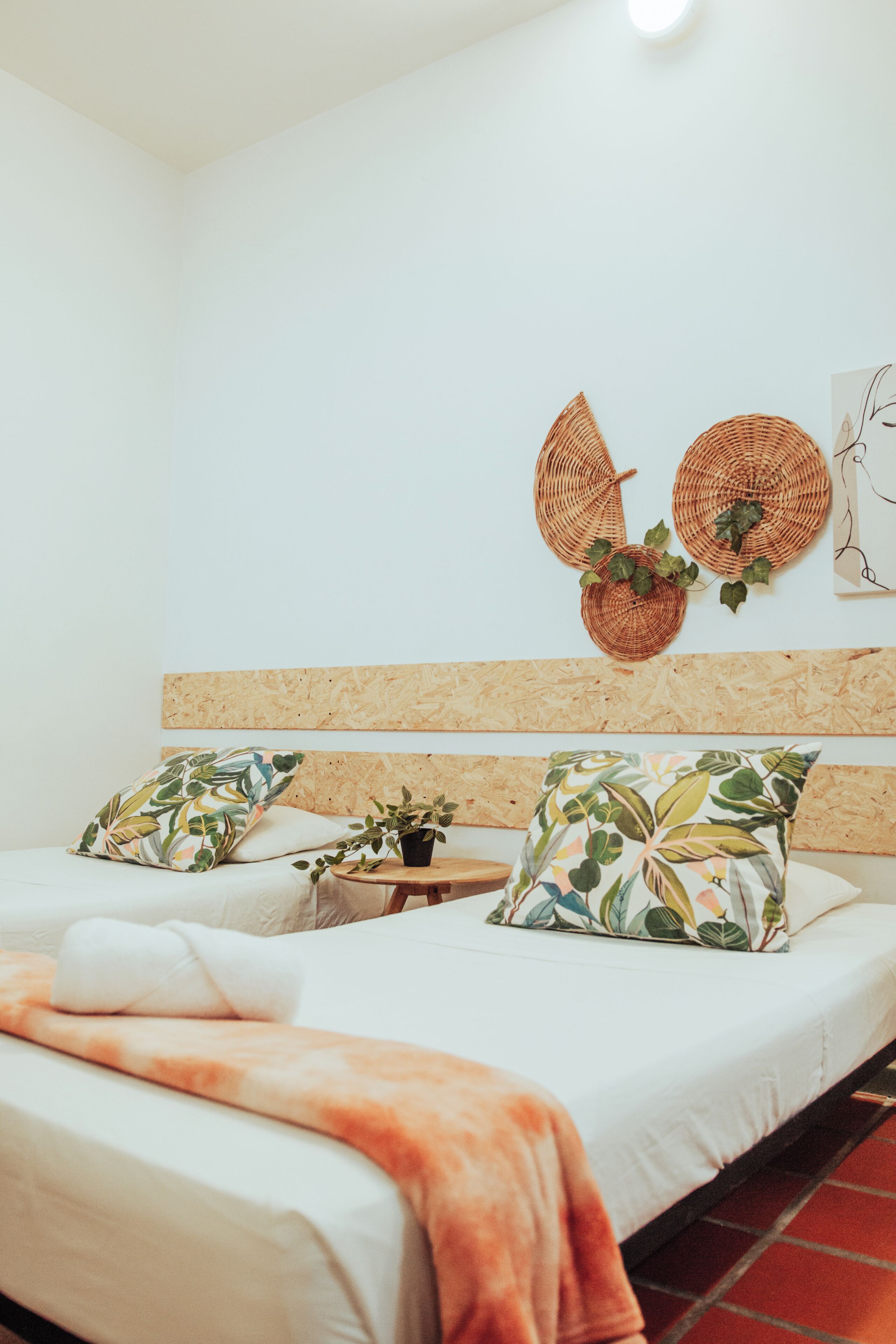 A room with a bed, two pillows, a blanket, a small table with a plant, and two wall-mounted baskets with greenery.
