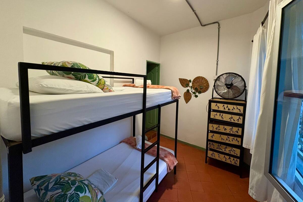 Quarto em dormitório com beliches, ventilador, armário fixado na parede e janela com cortinas.