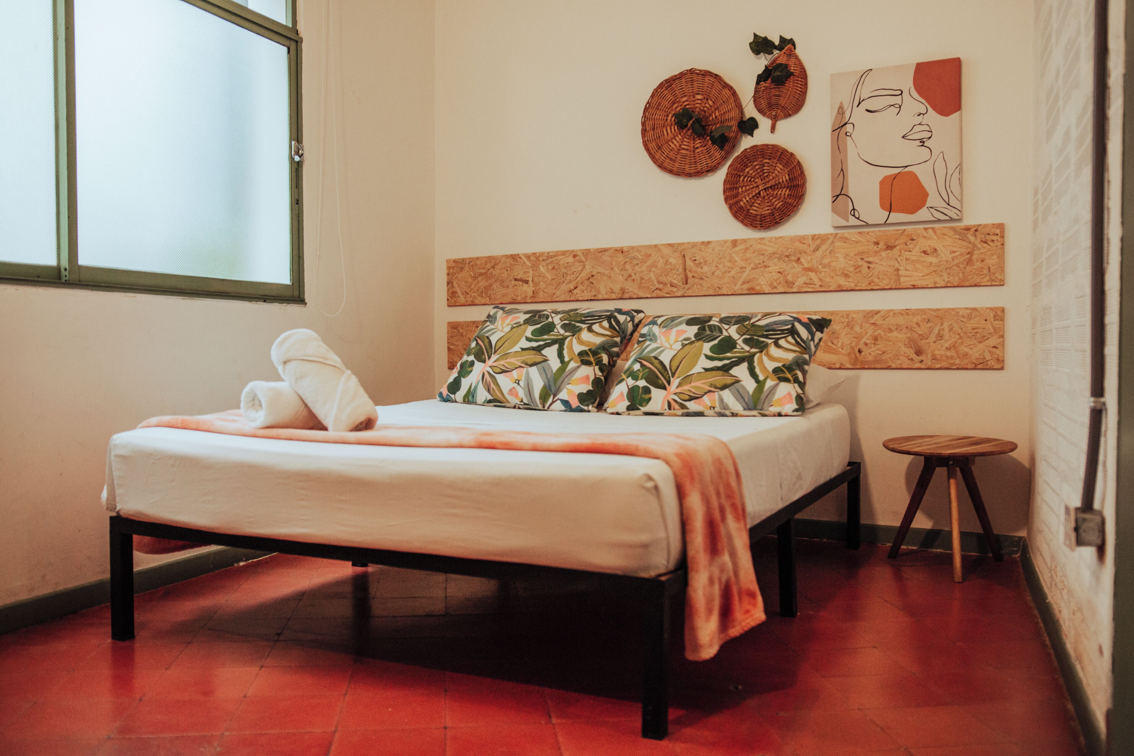 A bed with a white mattress and orange blanket, a small round wooden stool, and a corkboard wall with artwork.