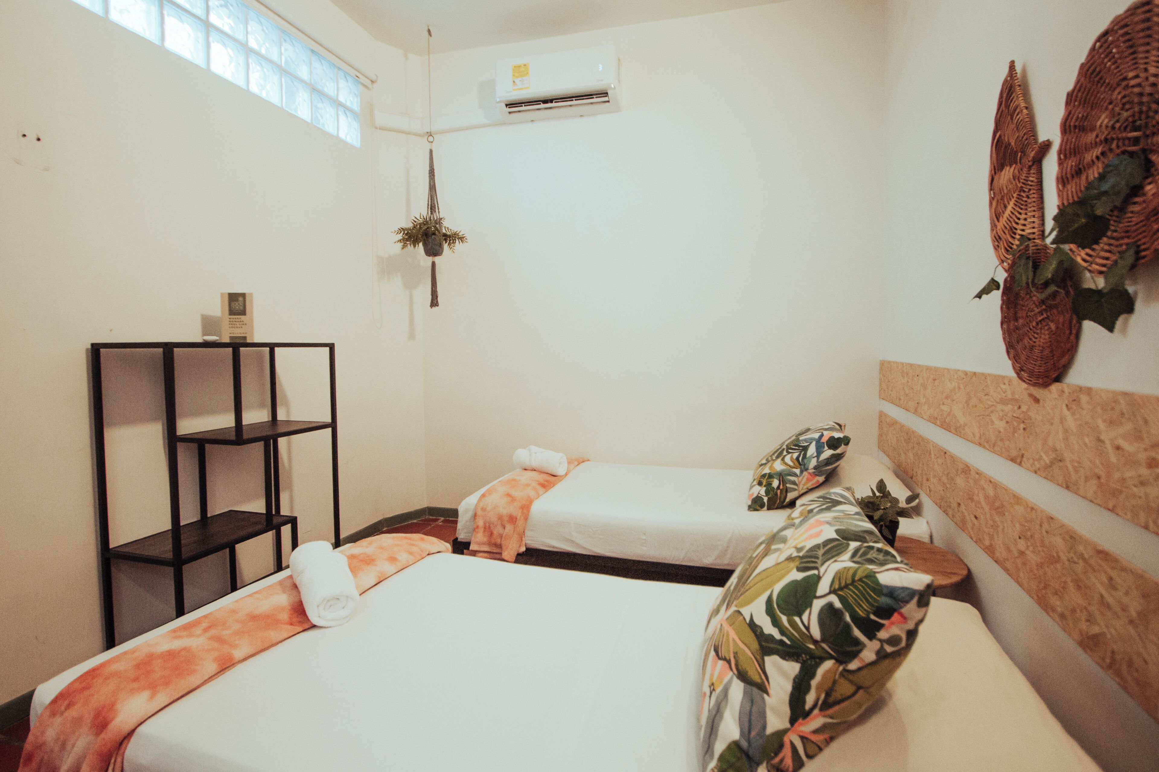 A hotel room with two beds, a shelf unit, an air conditioning unit, and a window.