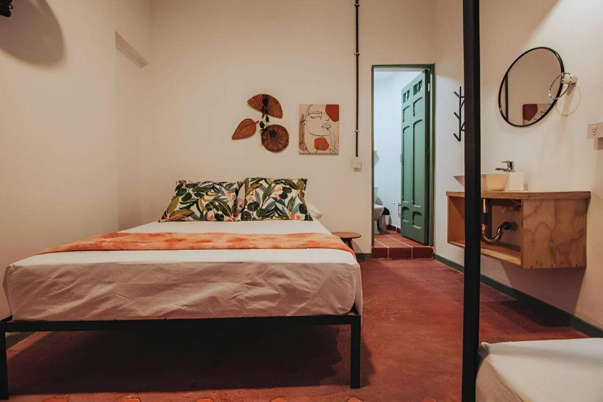 Quarto com cama, cómoda de madeira, espelho e porta que dá para outro espaço.