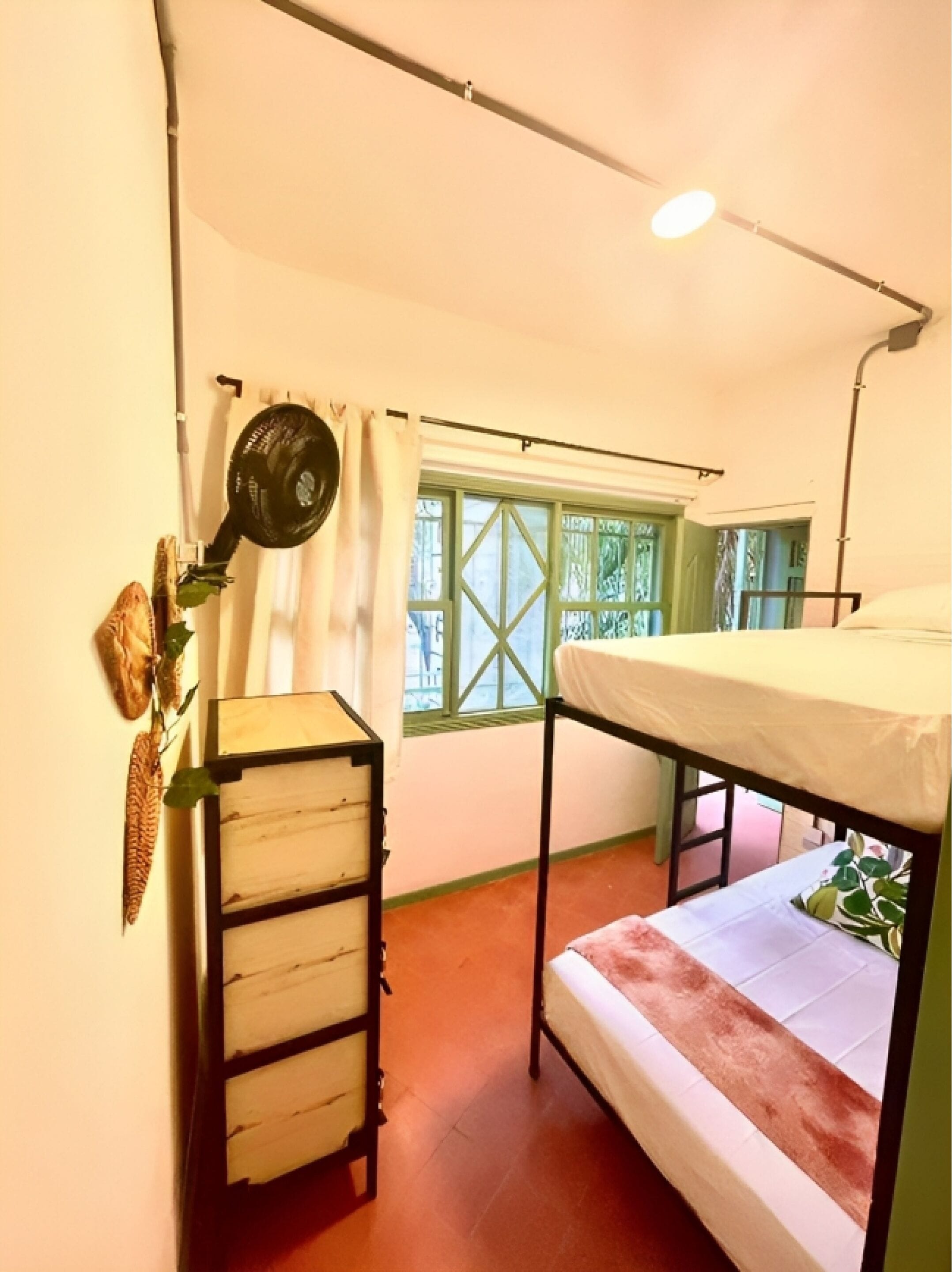 A small, single-room accommodation with a bunk bed, a wooden dresser, a ceiling fan, and a window with green shutters.