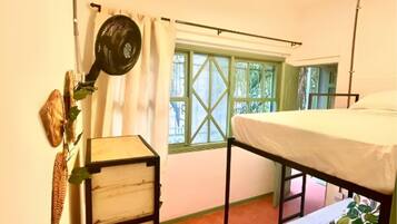 A small, single-room accommodation with a bunk bed, a wooden dresser, a ceiling fan, and a window with green shutters.