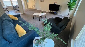 A living room with a gray carpet, a ceiling fan, a flat-screen TV, and a dining area with a table and chairs. - Comfy & spacious home (Austin)